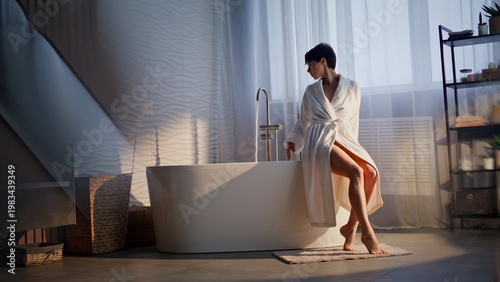 Tableau sur toile Serene brunette sitting bathtub checking water temperature soft sunlight room