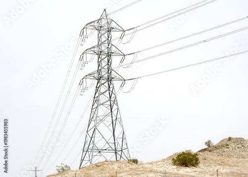 Large tall power lines running through desert landscape with mountains in the background. Hazy skies.