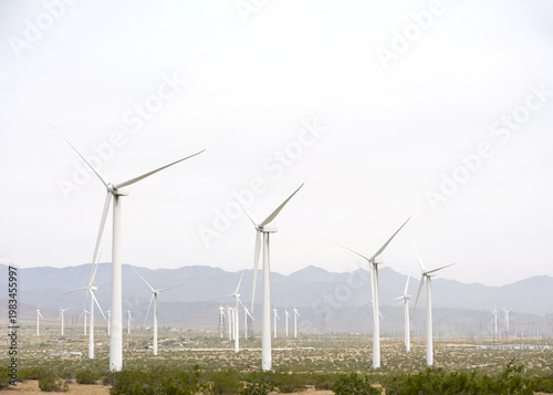 Desert landscape with windmills, low growing shrubs in the valley with mountains in background. Hazy sky with clouds.