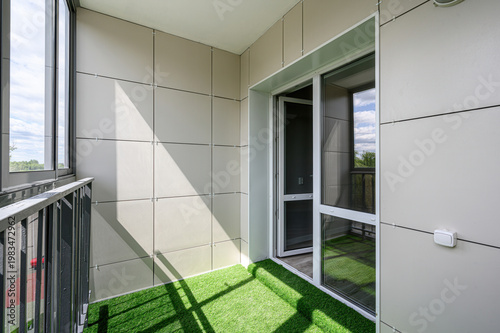 Bild auf Leinwand A modern balcony with beige panel walls, artificial green grass flooring, a glass sliding door, metal railing, and large windows offering a view of the sky and trees