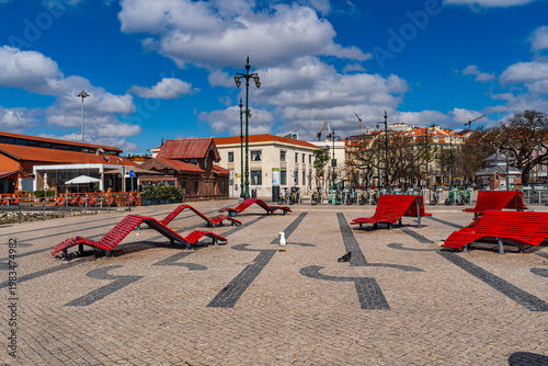 People relaxing on red benches in sunny urban square in Lisbon Portugal, vibrant city lifestyle scene with bicycles, pedestrians and European architecture
