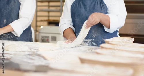 Female bakers, right baker stretching dough on floured bench readying boule for proofing baskets