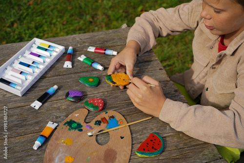 Boy applying paint to flat stones to create vegetable and fruit artwork on a table workspace.
