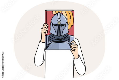 Book with knight on cover in hands of man covering face, reading literature about medieval soldiers. Man pretends to be knight in armor, dreaming of participating in gladiatorial tournaments.
