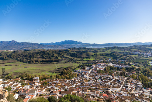 Aerial panoramic view of historic white town with tiled rooftops, located among green hills, farmland, and natural landscape. Cadiz province, Spain.