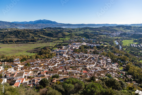 Aerial panoramic view of historic white town with tiled rooftops, located among green hills, farmland, and natural landscape. Cadiz province, Spain.