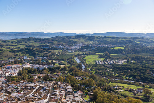 Aerial panoramic view of historic white town with tiled rooftops, located among green hills, farmland, and natural landscape. Cadiz province, Spain.