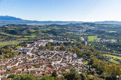 Aerial panoramic view of historic white town with tiled rooftops, located among green hills, farmland, and natural landscape. Cadiz province, Spain.