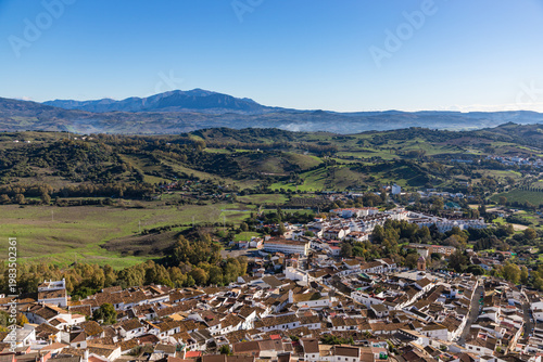 Aerial panoramic view of historic white town with tiled rooftops, located among green hills, farmland, and natural landscape. Cadiz province, Spain.
