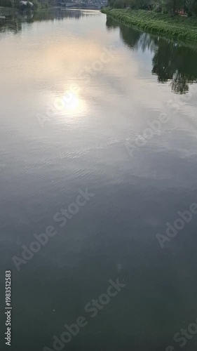 A calm river reflecting the sun at dawn or dusk. The calm water surface is rippled, with green banks and a bridge in the distance. Nature video.