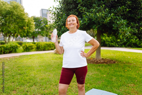 Fotografie Elderly woman in athletic clothing drinks water from bottle in a sunny park