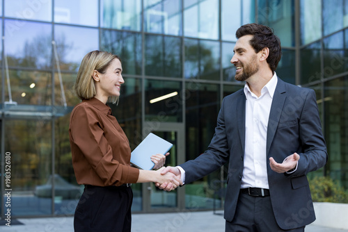 Two smiling business professionals are shaking hands, confirming a successful agreement and partnership outside a modern office building, symbolizing deal closing and collaboration