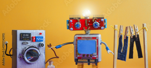 Foto Joyful home helper domestic assistant robot with red face and glowing head lamp holding power cord from toy washing machine, wooden drying rack with jeans and pants on clothesline