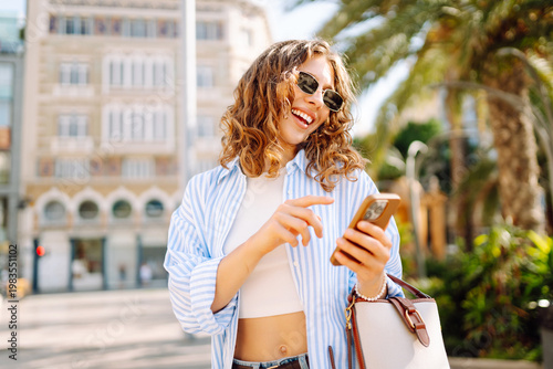 Fotografie Smiling woman stands on a city street with a phone among palm trees