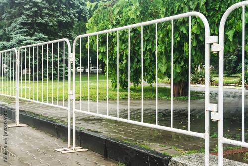 White metal pedestrian barriers standing along a wet sidewalk in a green urban park during rainy weather.