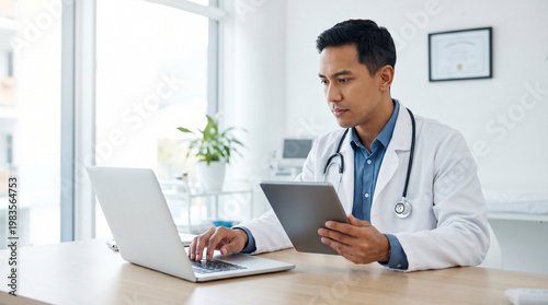 A male doctor, wearing a white coat and stethoscope, sits at a desk in a bright clinic. He uses both a laptop and a tablet, reviewing patient data with a focused, professional expression.