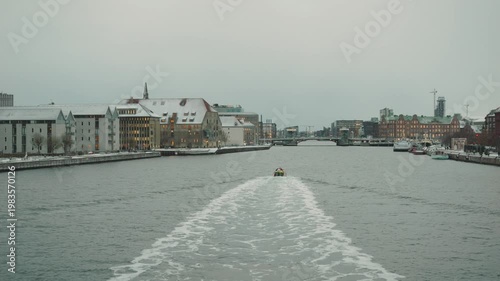 A tranquil river scene featuring a boat gliding through calm waters, surrounded by urban architecture and wintery ambiance. The image captures the peaceful interaction between nature and city life.