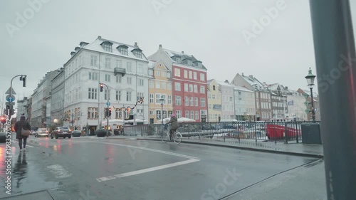 A vibrant scene of colorful buildings lining a waterfront on a rainy day, showcasing reflections in the wet pavement and a cyclist passing by, creating an inviting atmosphere.