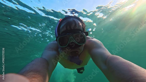 A man wearing a mask and snorkel swims underwater in the ocean, exploring the underwater world and filming himself on camera