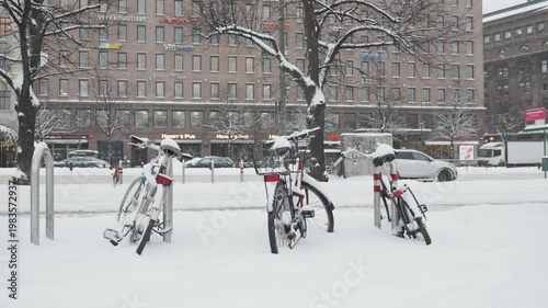 A scene depicting bicycles blanketed in snow against a backdrop of a bustling urban environment, showcasing the contrast between nature's beauty and city life during winter.