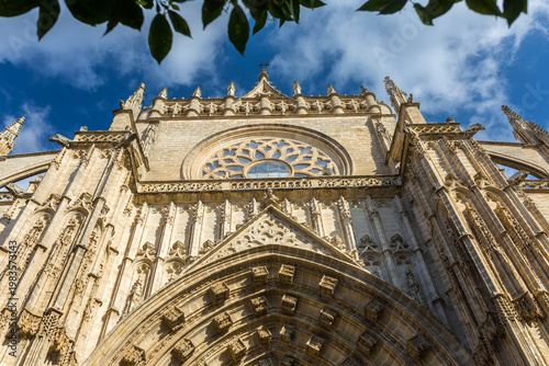 Seville, Andalusia, Spain: magnificent gothic facade of the historic cathedral of seville with intricate stone carvings, ornate rose window, and flying buttresses under a clear blue sky