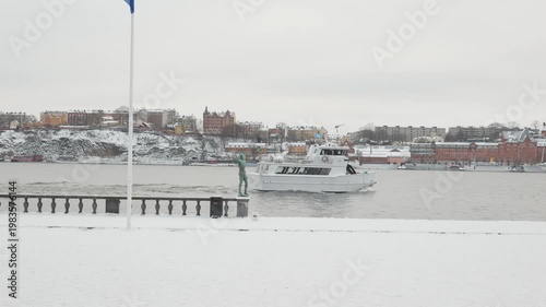 A picturesque winter scene featuring a boat cruising through icy waters, surrounded by snow-covered buildings and a tranquil cityscape, evoking a serene and chilly atmosphere.