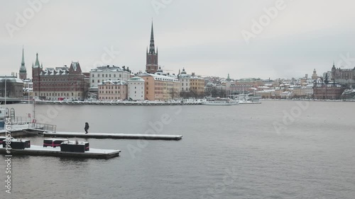 A serene winter scene featuring a snowy cityscape with historical buildings and a harbor. The landscape captures the tranquility of a cold day, showcasing boats in the water and soft white snow blanke