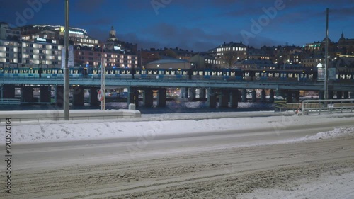 A serene cityscape featuring snow-covered areas and soft reflections on the water. The scene showcases illuminated buildings and architecture against a darkening sky, creating a tranquil winter atmosp