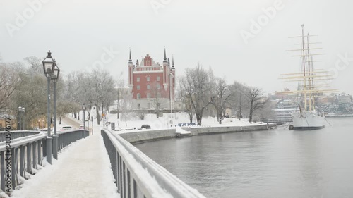A serene winter scene featuring a snow-covered riverbank lined with trees and a grand historic building in the background. A ship rests calmly on the water, creating a picturesque landscape.