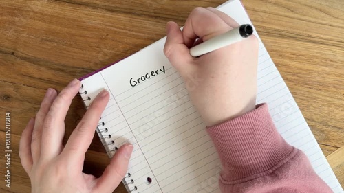 Close up of hands writing “grocery list” on lined notebook paper, everyday shopping and meal planning concept, natural light overhead shot on wooden table, 4K video