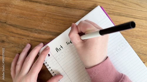 Close up of hands writing “art supplies needed” on lined notebook paper, creative planning and shopping list concept for artists, natural light overhead shot on wooden table, 4K video