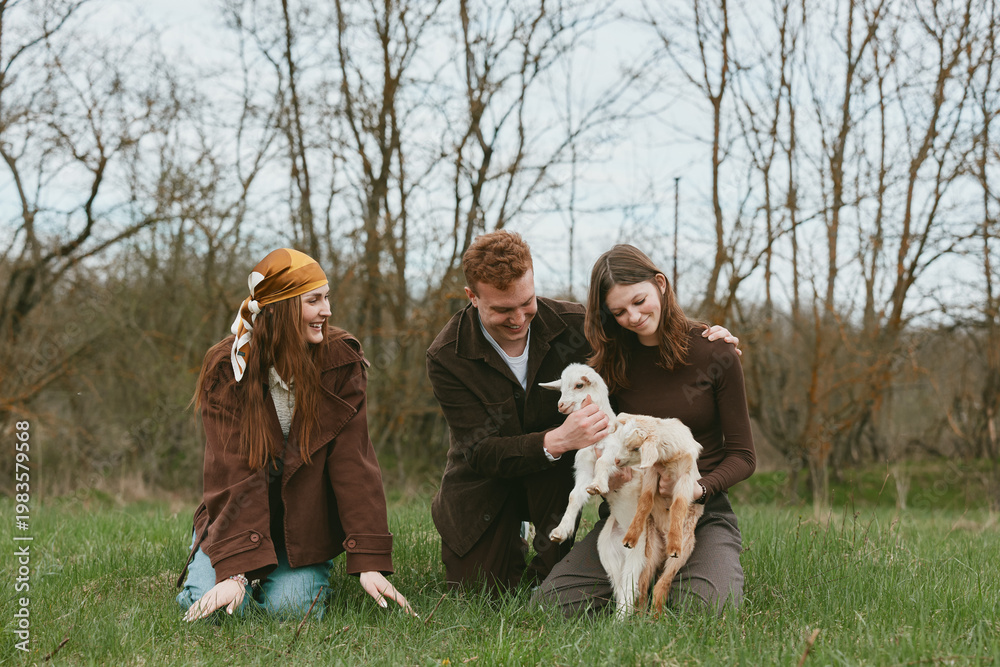 Fototapeta premium Cheerful group outdoors with a young goat kid, friends sharing a gentle moment on lush grass, candid interaction, natural light, relaxed mood, countryside setting