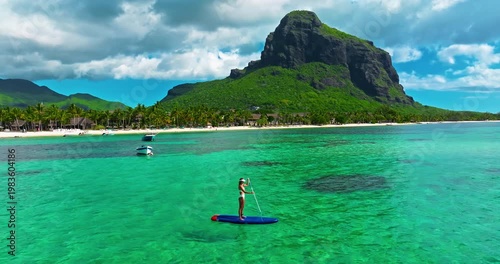 Aerial view of woman in white swimsuite standing on sup board with paddle on crystal turquoise water with coral reef on the tropical island in Indian ocean. Active water sport. Mauritius island