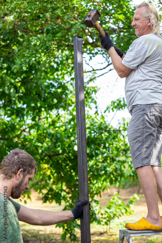 Two men working together to install a metal fence post in a garden. Teamwork, construction site, manual labor, home improvement.
