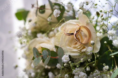 Two gold wedding rings on a bridal bouquet with white roses and gypsophila. Elegant marriage symbols and romantic floral background for engagement and wedding concepts, selective focus