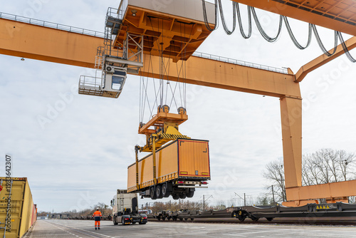 Gantry crane lifting a yellow semi-trailer onto a freight train at an intermodal terminal, logistics worker overseeing cargo loading for rail transport and combined transportation