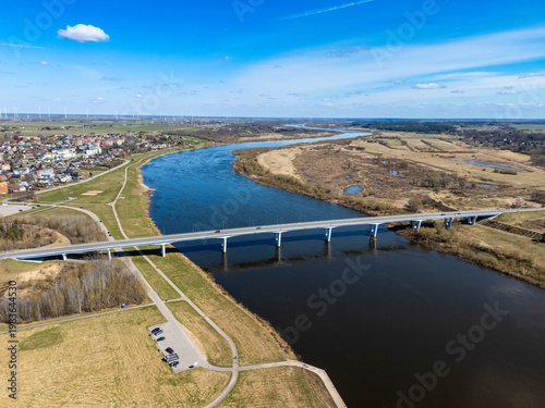 Aerial drone view of a modern bridge crossing a wide Nemunas river in a rural landscape with fields and small town in the distance under blue sky.