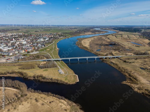 Aerial drone view of a modern bridge crossing a wide Nemunas river in a rural landscape with fields and small town in the distance under blue sky.