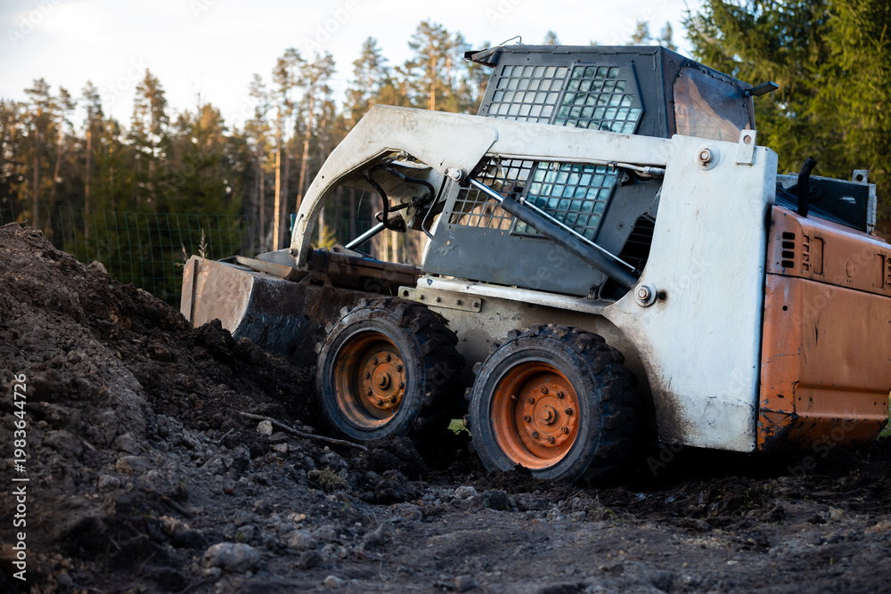 Fototapeta premium White and orange skid steer climbs a muddy slope, cab grid visible, thick tread tires caked with soil. Conifers surround cleared earth in daylight, low angle three quarter view.