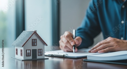 Person signing real estate contract with house model, representing home purchase, property investment, mortgage agreement and financial approval.