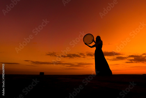 Silhouette of woman playing Erbane (frame drum) on rooftop at sunset in Mardin, Türkiye, with dramatic orange sky reflecting cultural traditions and artistic expression