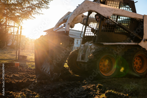 Compact skid steer with orange wheels digs wet soil near a metal fence and masonry, bucket raised as sunburst flares through cage grilles, low angle shows chunky treads.