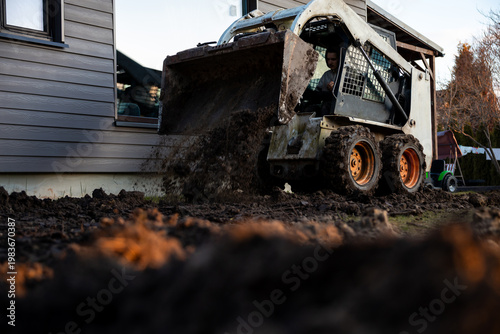 A compact skid steer dumps wet soil against a house foundation with gray siding. Late afternoon light highlights flying clods, muddy tires, ruts, and motion blur at ground level.