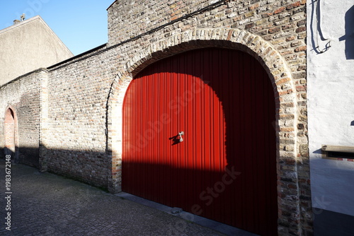 Garagentor in leuchtendem Rot mit Rundbogen in altem Mauerwerk aus Bruchstein und Naturstein bei blauem Himmel und Sonnenschein in den Gassen der Altstadt von Brügge in Westflandern in Belgien