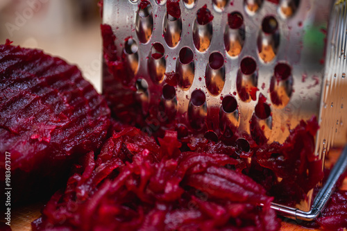 Macro of boiled red beetroot being grated on a metal grater on a wooden board. Healthy cooking process for salad or borscht. Vibrant magenta vegetable food texture