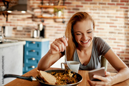 Smiling woman eating breakfast while using smartphone at home kitchen