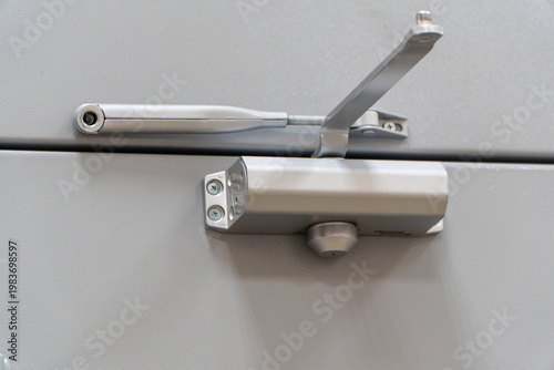 silver hydraulic automatic door closer mechanism with adjustable metal arm installed on top of gray fireproof entrance door in commercial building.