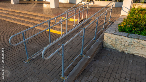 Photos A concrete accessibility ramp featuring dual grey metal handrails leading to a building entrance, with stone-paved steps and a flower bed on the side under warm late afternoon sunlight