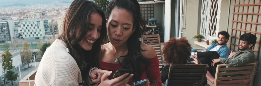 Obraz premium Girls chatting, using mobile phone while standing on balcony on modern city on the background. Panoramic