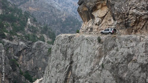 Panoramic view shows a 4x4 vehicle driving along a dangerous road on a very high rocky cliff. Offroad travel clings to the steep edge as dramatic terrain drops into the abyss.